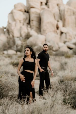 Couple in black attire walking through desert landscape with dramatic rock formations.
