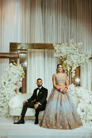 Elegant couple in traditional South Asian wedding attire pose with white floral decorations and sheer curtain backdrop.