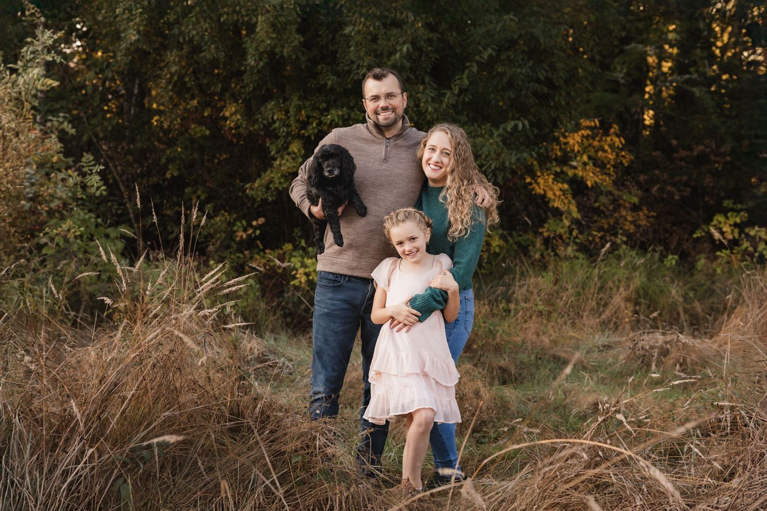 A family and their black dog pose together in a fall outdoor setting with golden grass and autumn trees in the background.