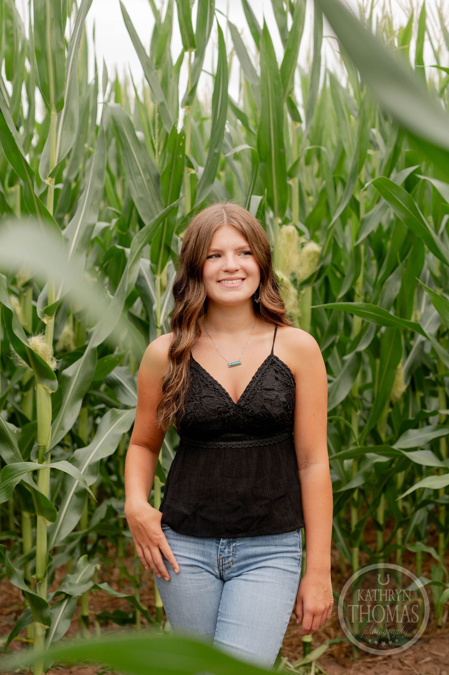 Casual portrait of woman in black tank top standing among tall green corn stalks.