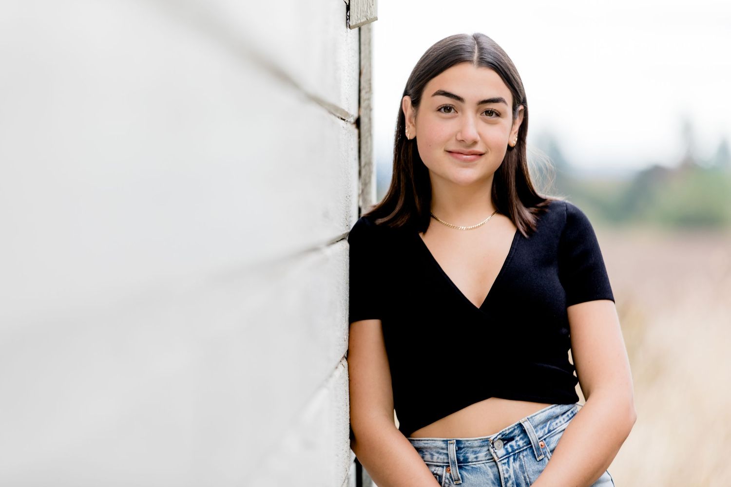 A person in a black crop top and denim jeans leans against a white wall outdoors.