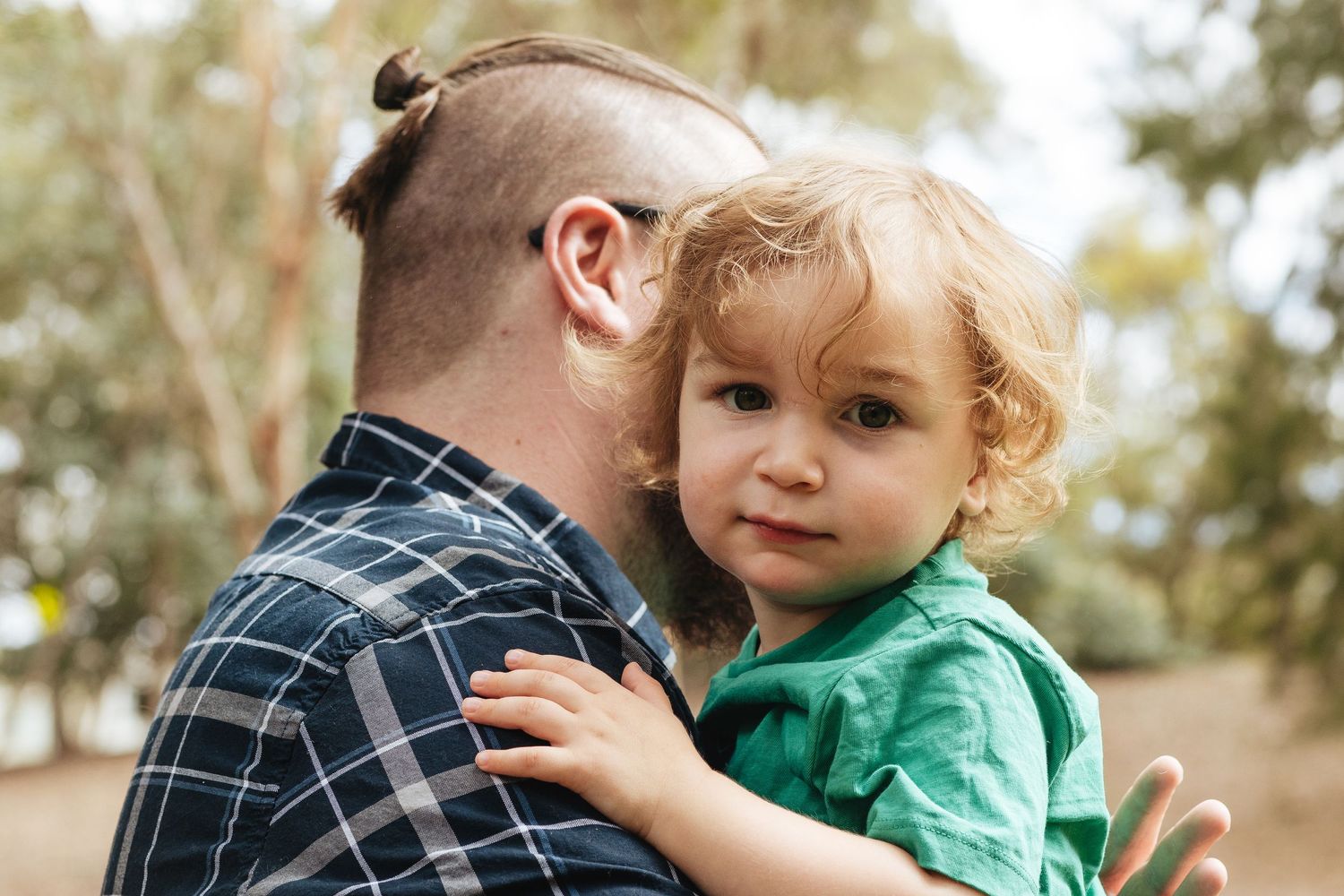 A young child wearing a green shirt rests on someone's shoulder while being held outdoors.
