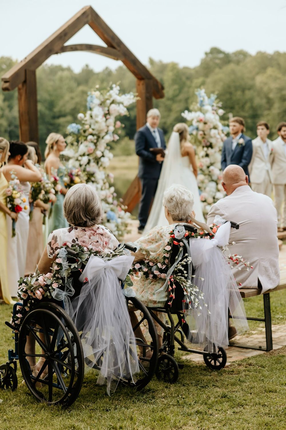 An outdoor wedding ceremony under a wooden arch with decorative floral arrangements and wheelchairs decorated in white tulle.