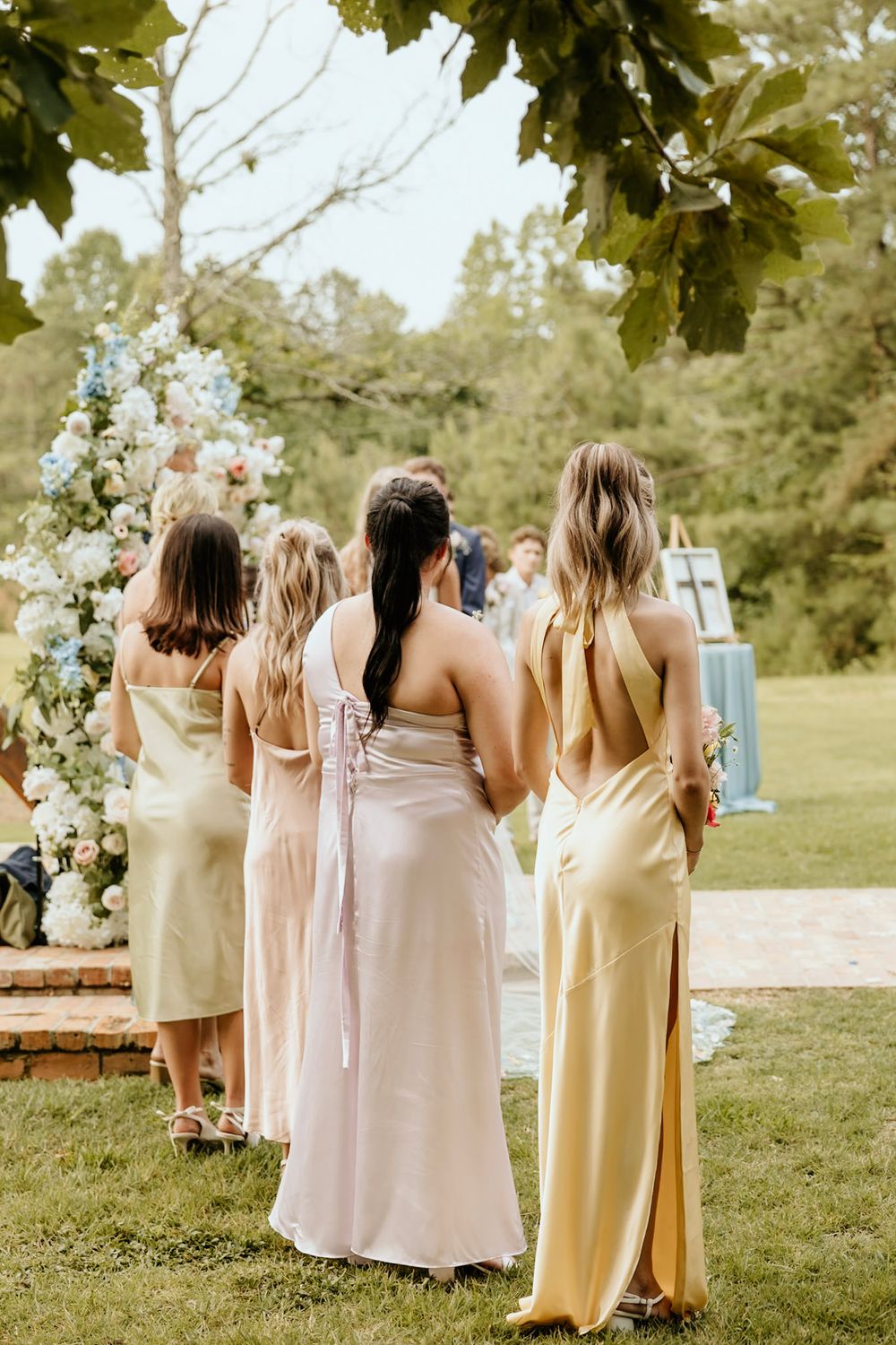 Bridesmaids wearing pastel dresses stand together viewing a floral arch at an outdoor wedding ceremony.