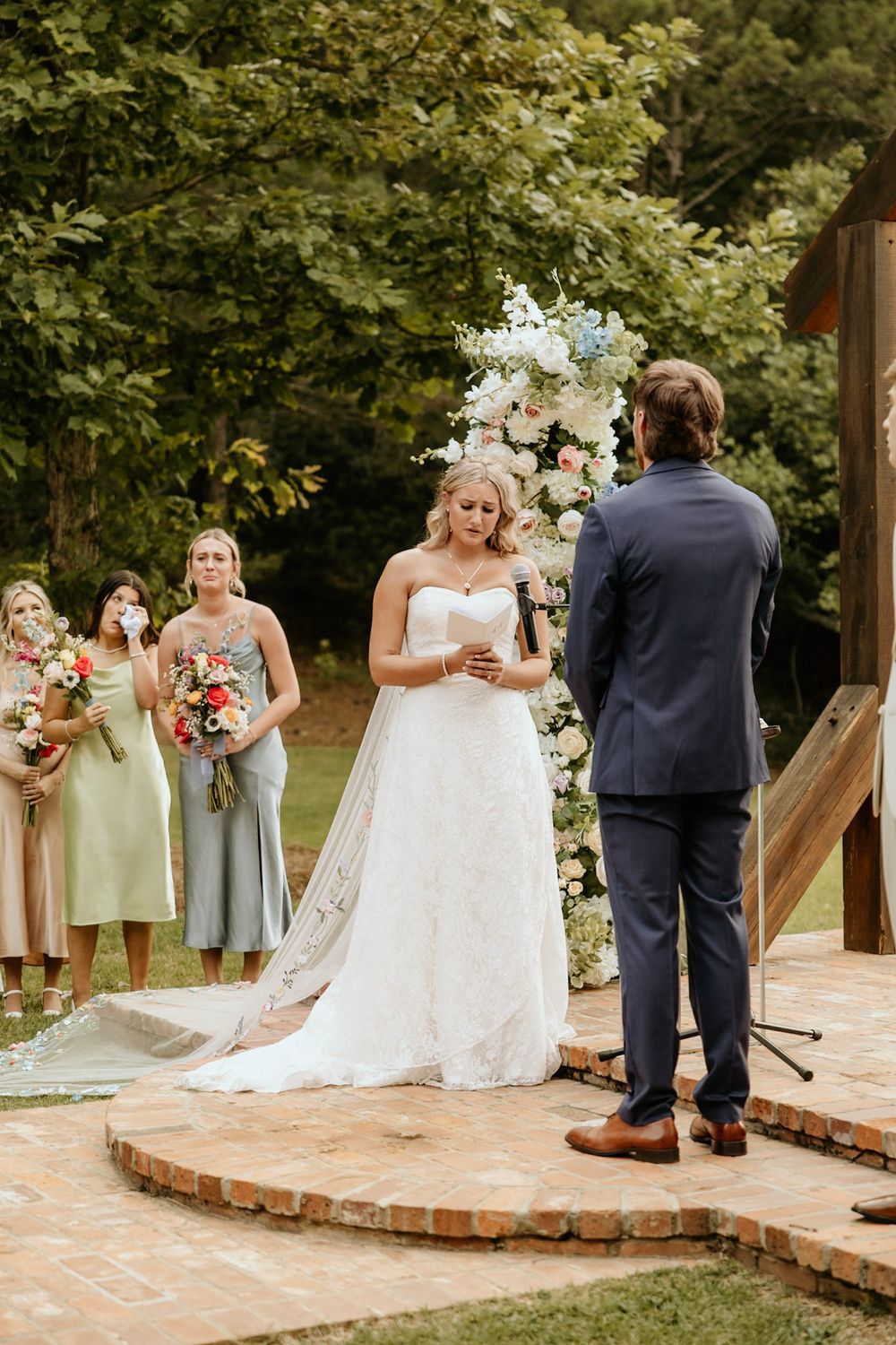 A wedding ceremony takes place on a brick platform with an elegant white floral arch in a garden setting.