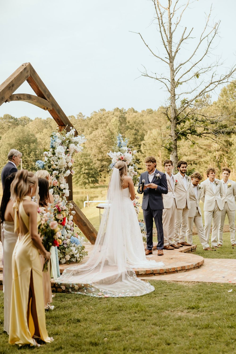 Outdoor wedding ceremony at rustic wooden arch decorated with floral arrangements and greenery in natural setting.