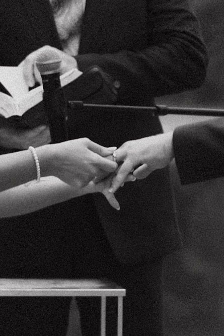 A close-up black and white photograph shows two hands exchanging a ring during an intimate wedding ceremony moment.