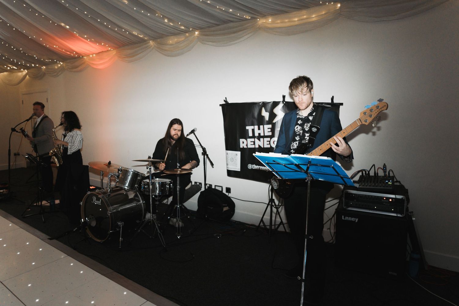 Band performs on stage with drummer, bass player and string lights decorating the ceiling at an indoor venue.