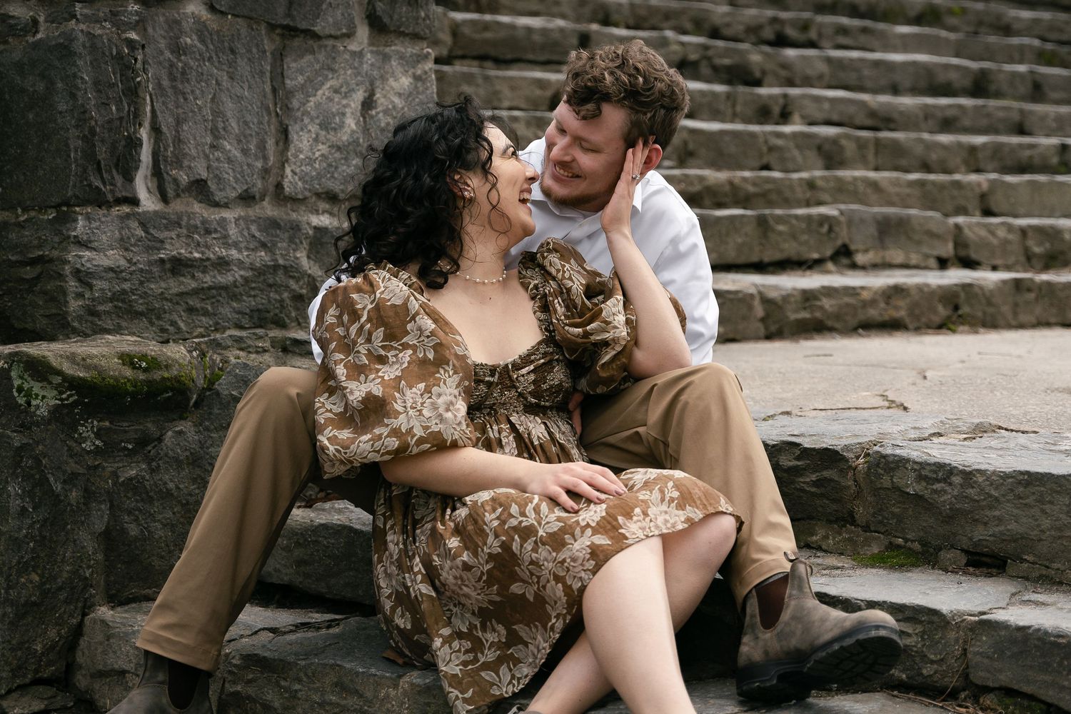 A romantic couple shares an intimate moment while sitting on stone steps, the person in brown floral dress laughing joyfully.