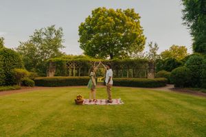 A couple enjoys a romantic picnic on a manicured lawn surrounded by lush garden hedges and trees.