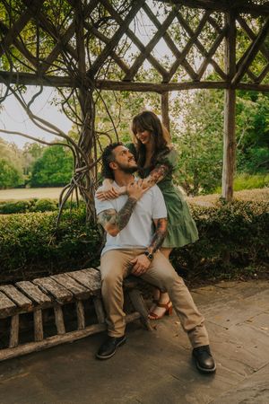 A couple shares a tender moment under a wooden garden pergola covered in vines on a sunny afternoon.