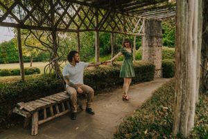 Two people share a moment in a lush garden pergola with latticed overhead beams during a sunny afternoon.
