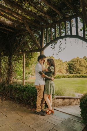 A couple shares an intimate moment under a vine-covered pergola in a garden setting.
