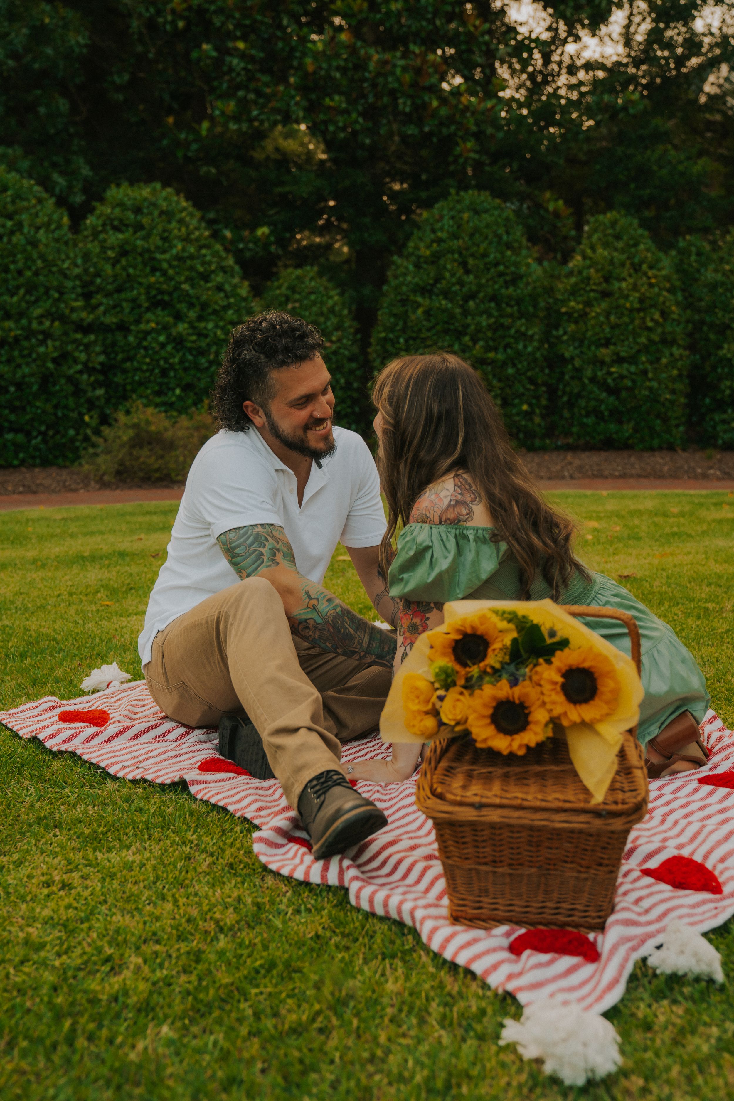 Couple enjoying romantic picnic on striped blanket with sunflowers in wicker basket on green lawn by garden.