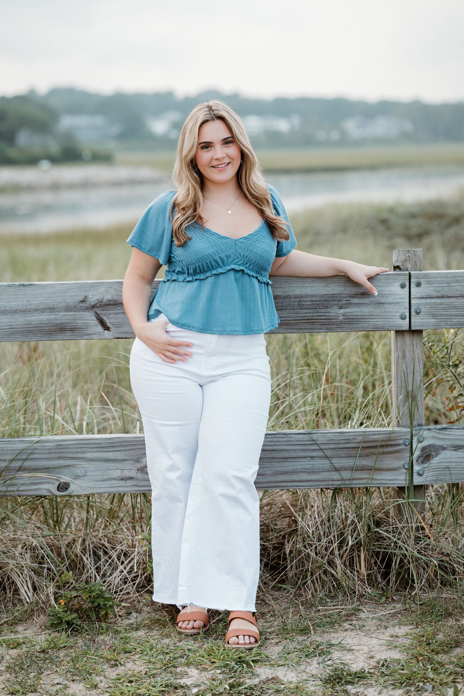 A person wearing a blue ruffled top and white pants leans against a wooden fence near a coastal marsh landscape.