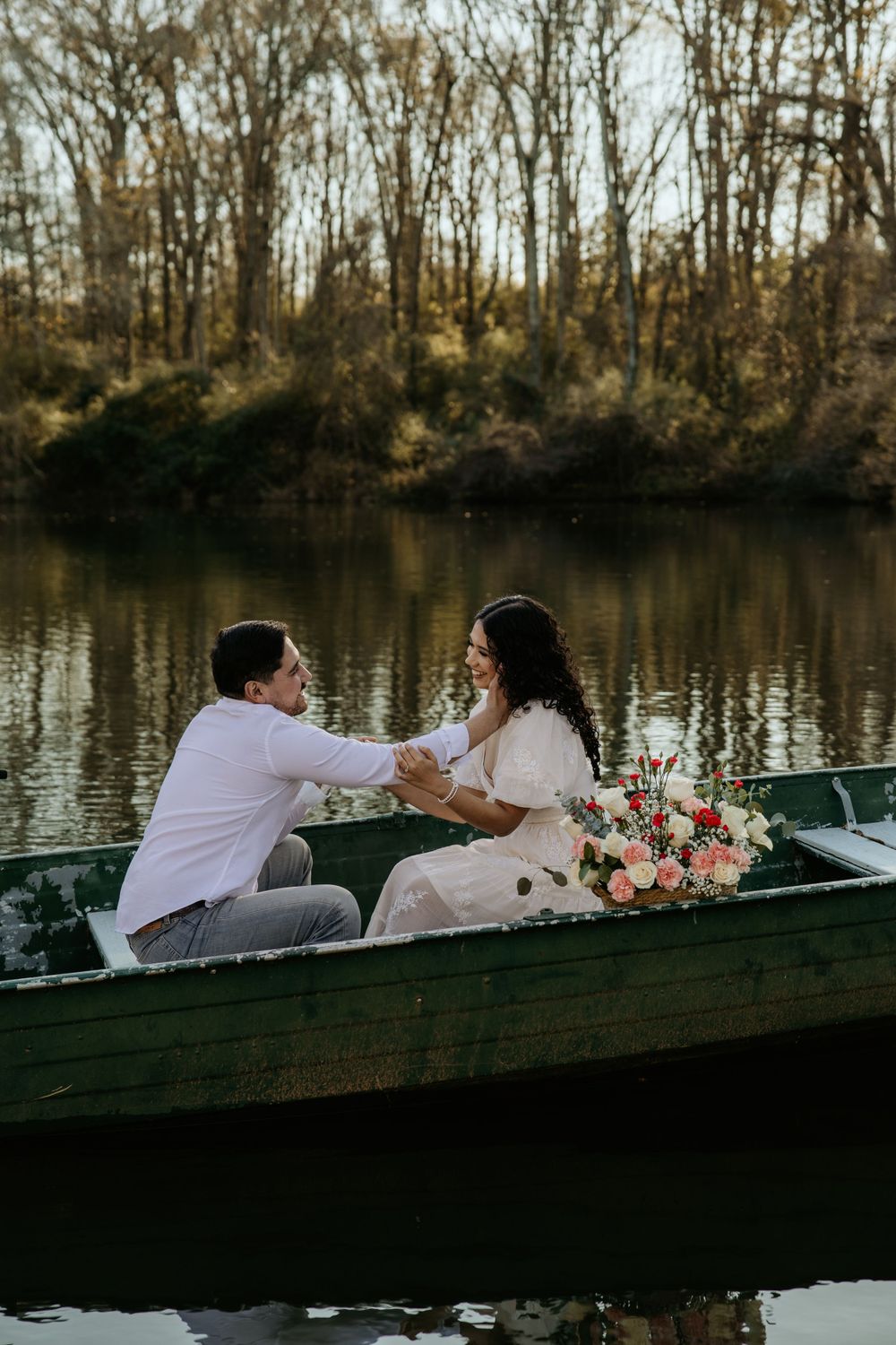 A romantic couple sits in a wooden boat on a serene lake with a bouquet of pink flowers at sunset.