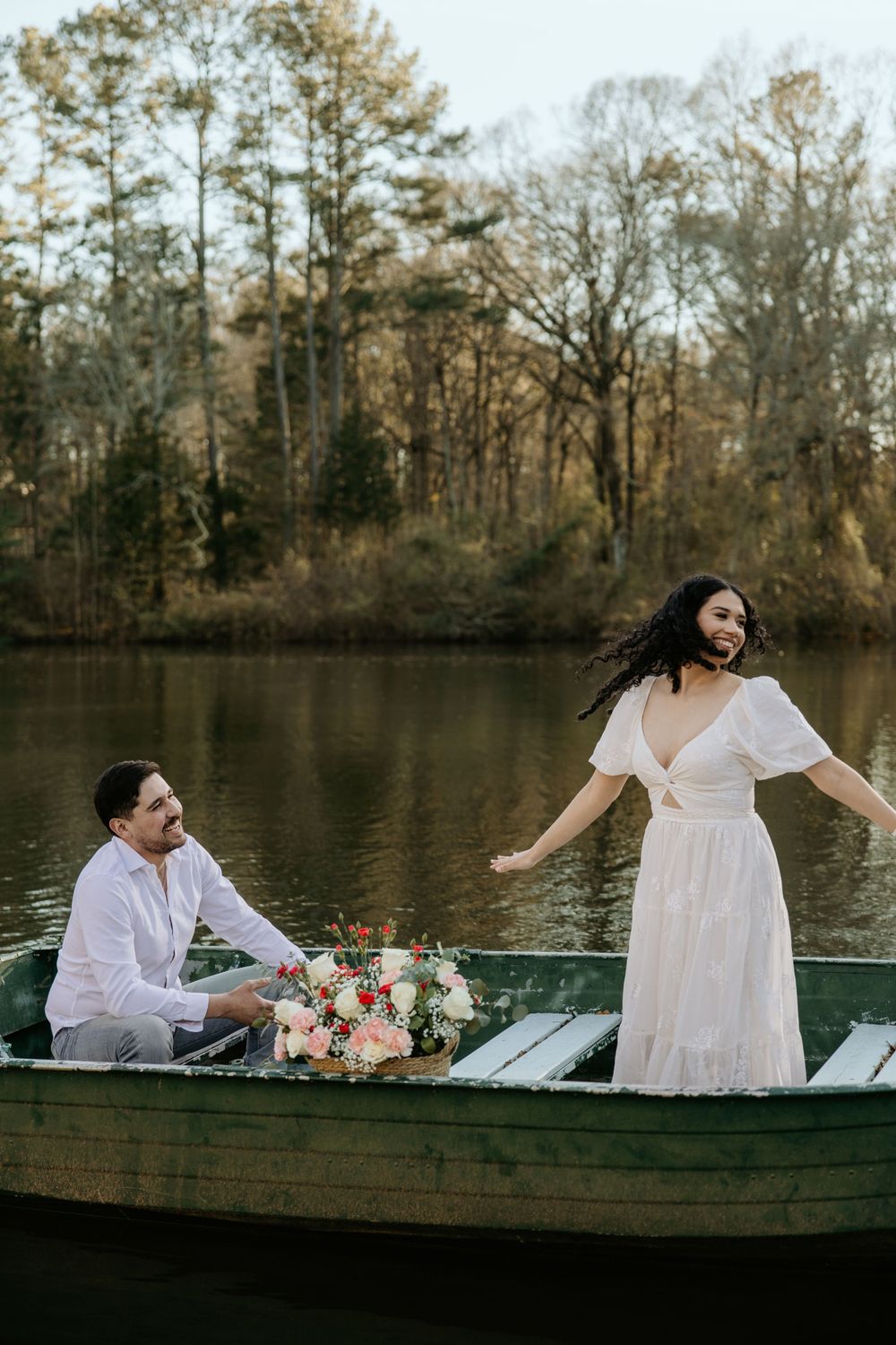 A romantic scene with two people in a rowboat on a lake surrounded by trees, with a bouquet of pink flowers.