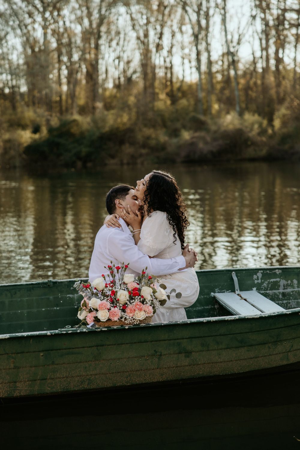 Romantic couple shares tender moment in green rowboat on lake with floral bouquet at sunset among bare trees.