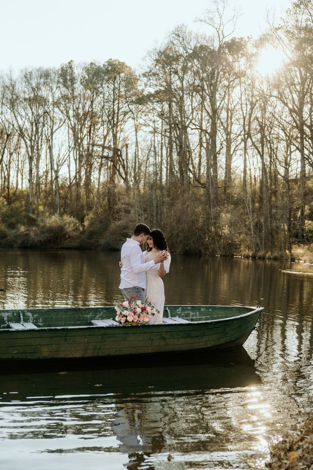 A couple shares an intimate moment while standing in a wooden boat on a tranquil lake surrounded by bare trees.