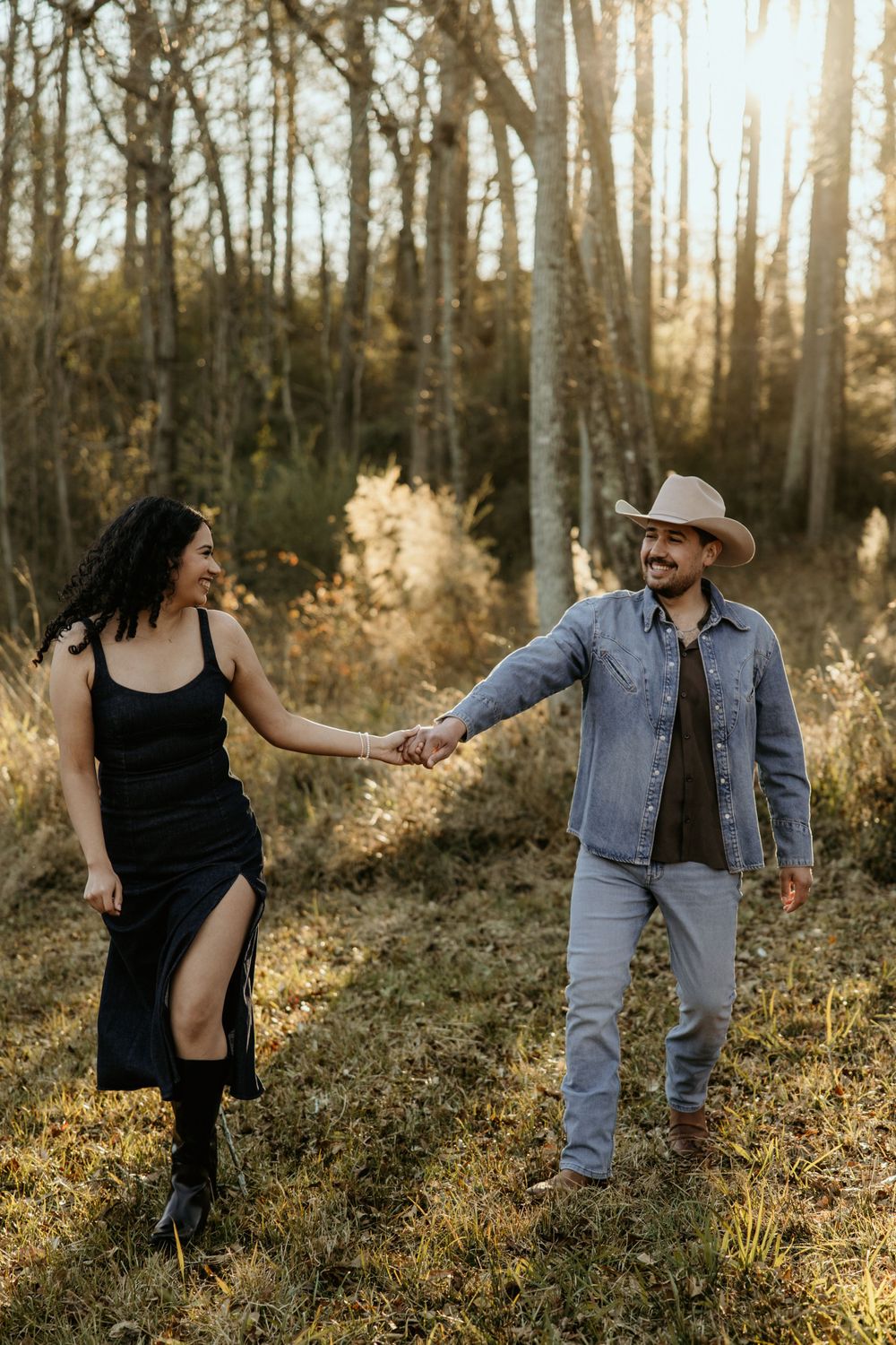 A couple holds hands in a sunlit forest clearing during golden hour, with her wearing a black dress and him in a cowboy hat.