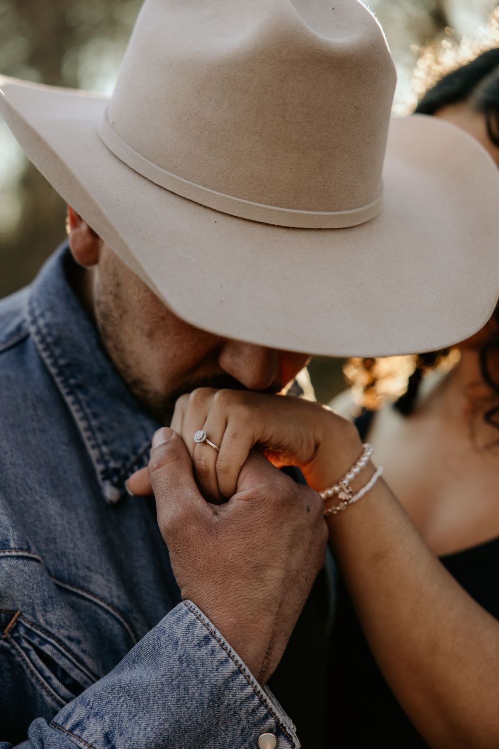 Close-up of clasped hands showing engagement rings, with person wearing denim jacket and light beige cowboy hat.