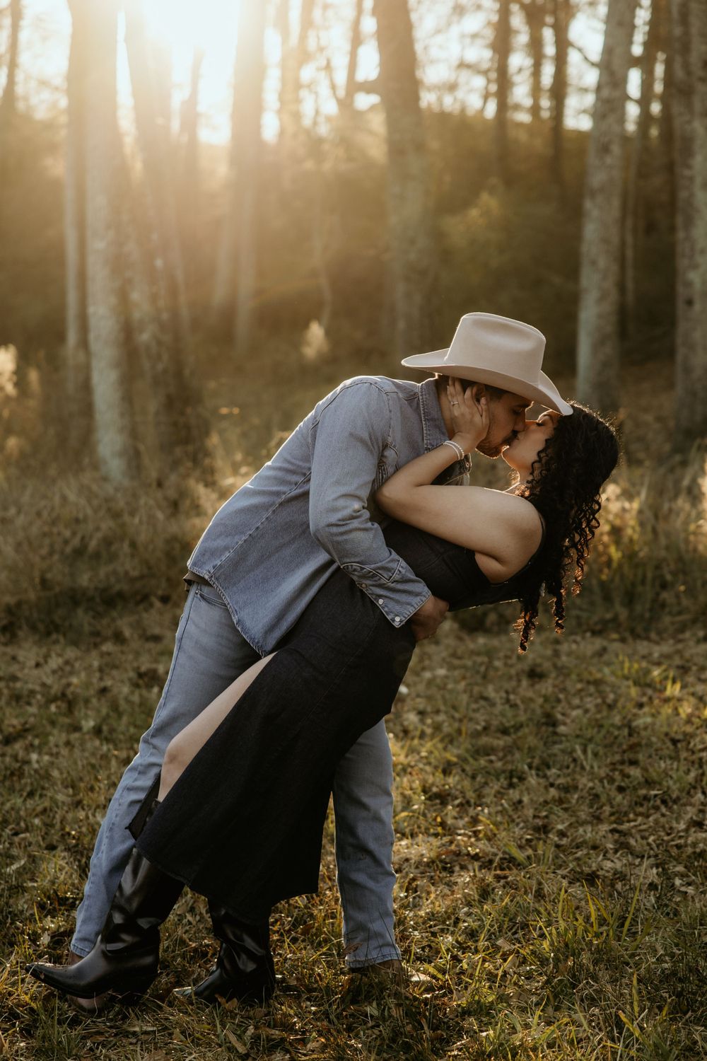Romantic couple sharing a passionate kiss in a sunlit forest at golden hour while wearing casual western attire.