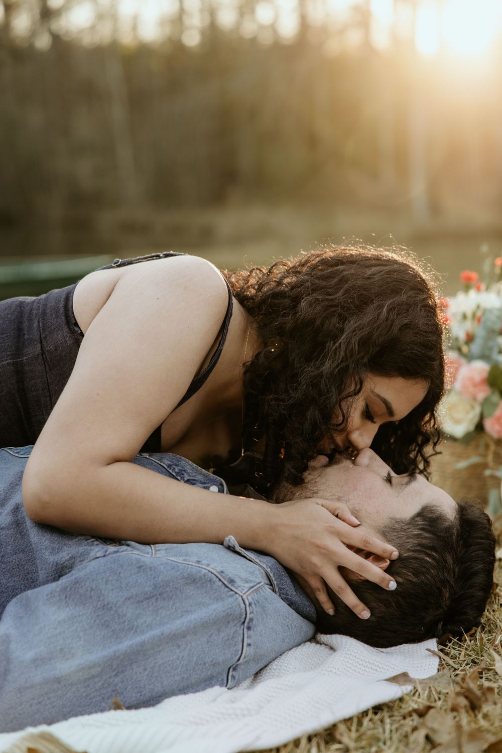Two people share an intimate moment while lying on a blanket outdoors as golden sunlight filters through trees behind them.