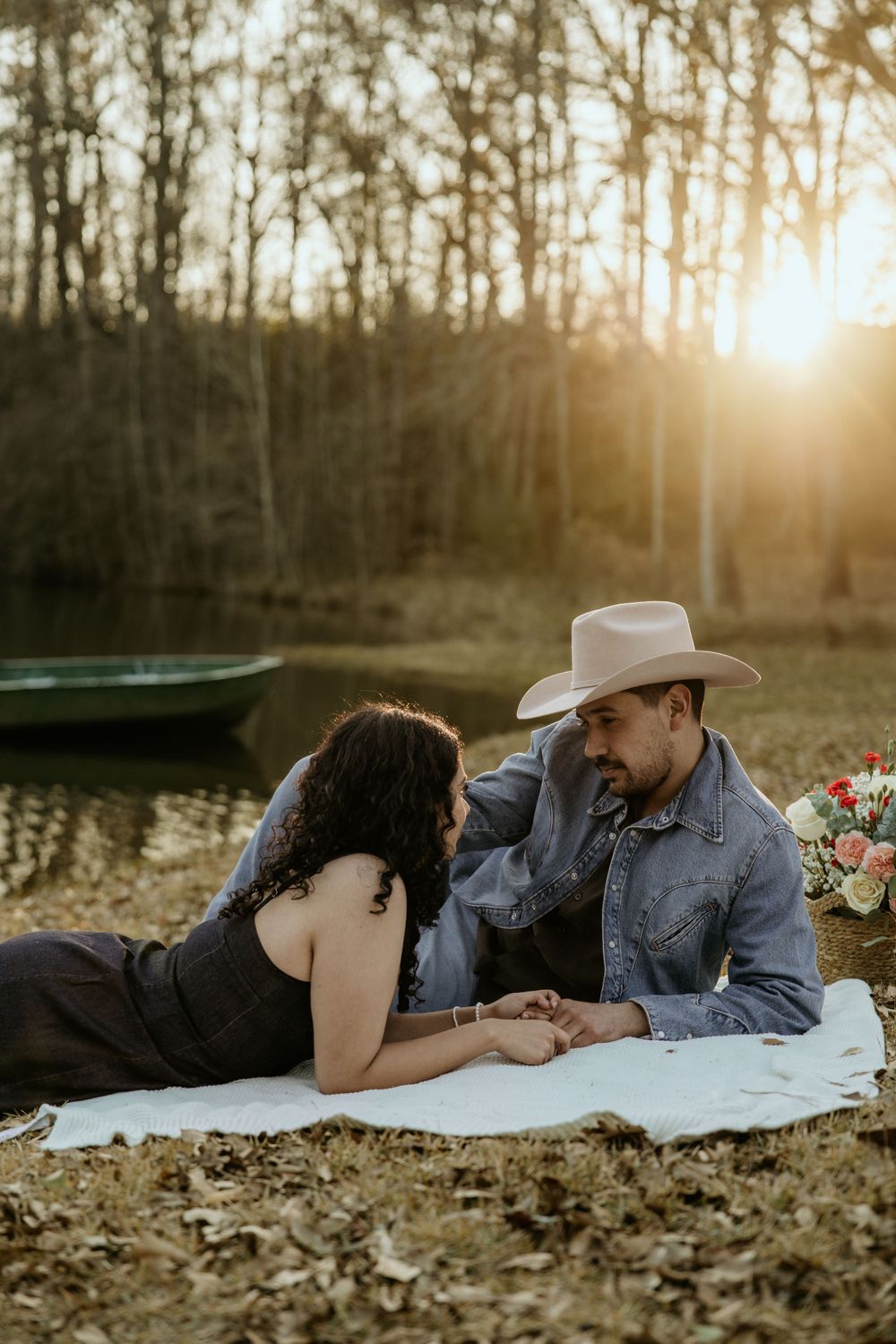 Two people share a romantic moment on a blanket at sunset near a pond with flowers and a cowboy hat.