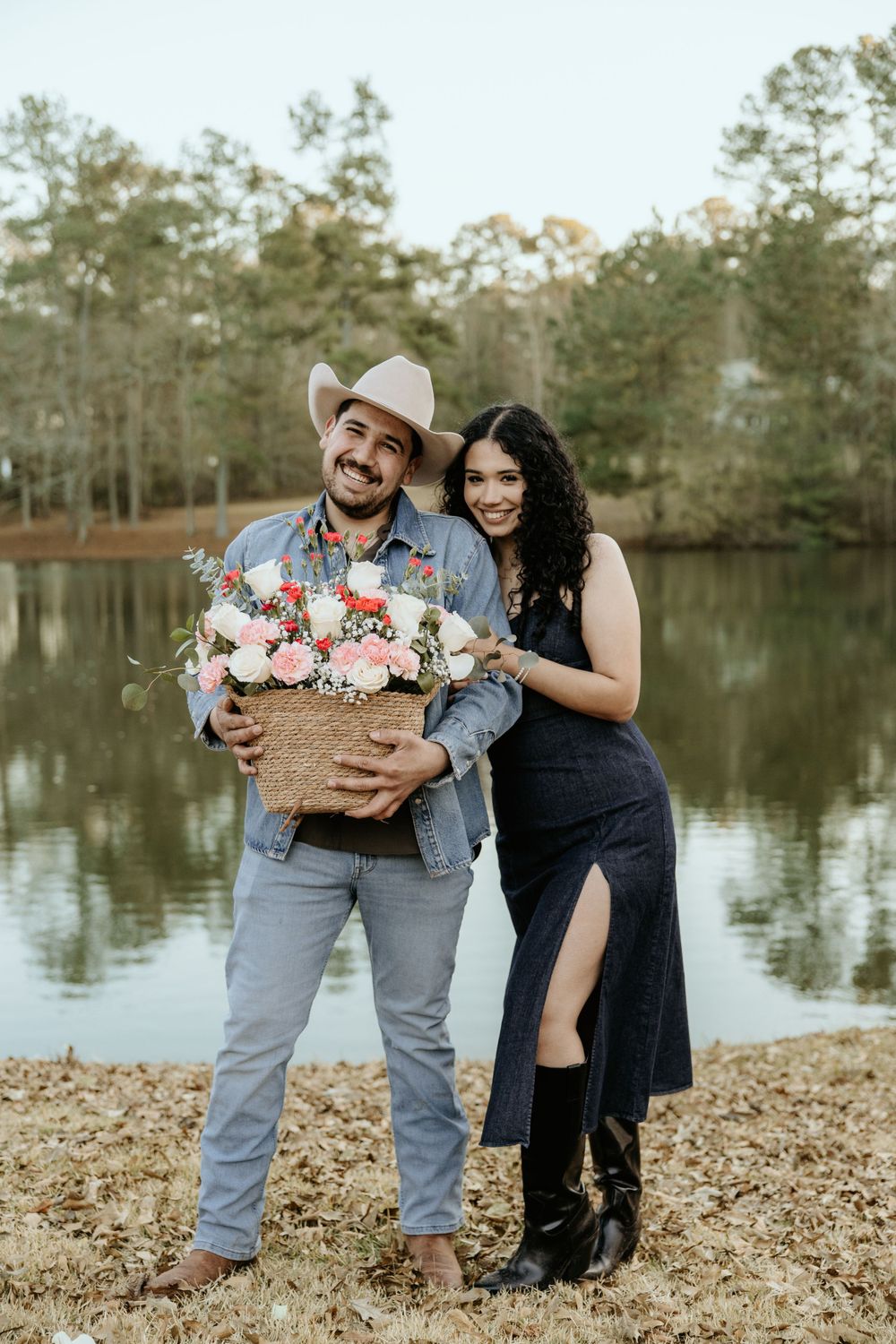 A couple poses with a large basket of flowers by a lake, with one person wearing a cowboy hat and boots.