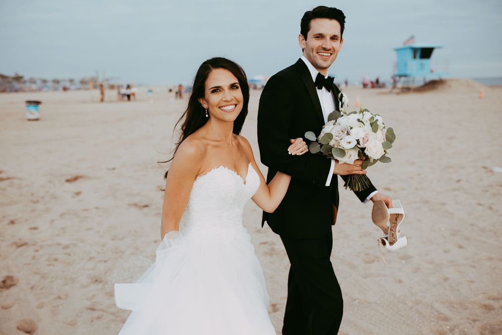 A couple in wedding attire walks and smiles on a sandy beach with lifeguard towers visible in the background in Orange County.