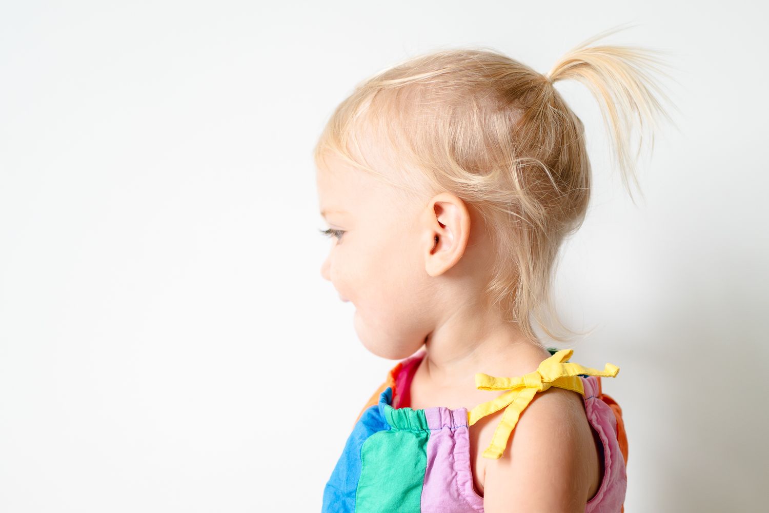 A young child with blonde hair in a ponytail wearing a colorful sleeveless dress looks to the side against a white background.