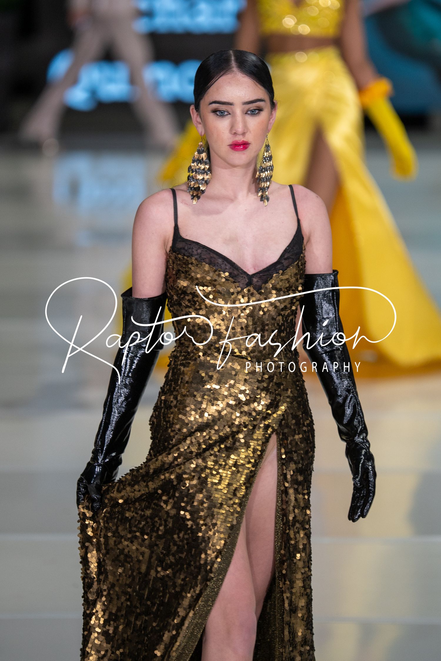 Model walks runway in sparkling gold evening gown with thigh slit and black gloves at fashion show.