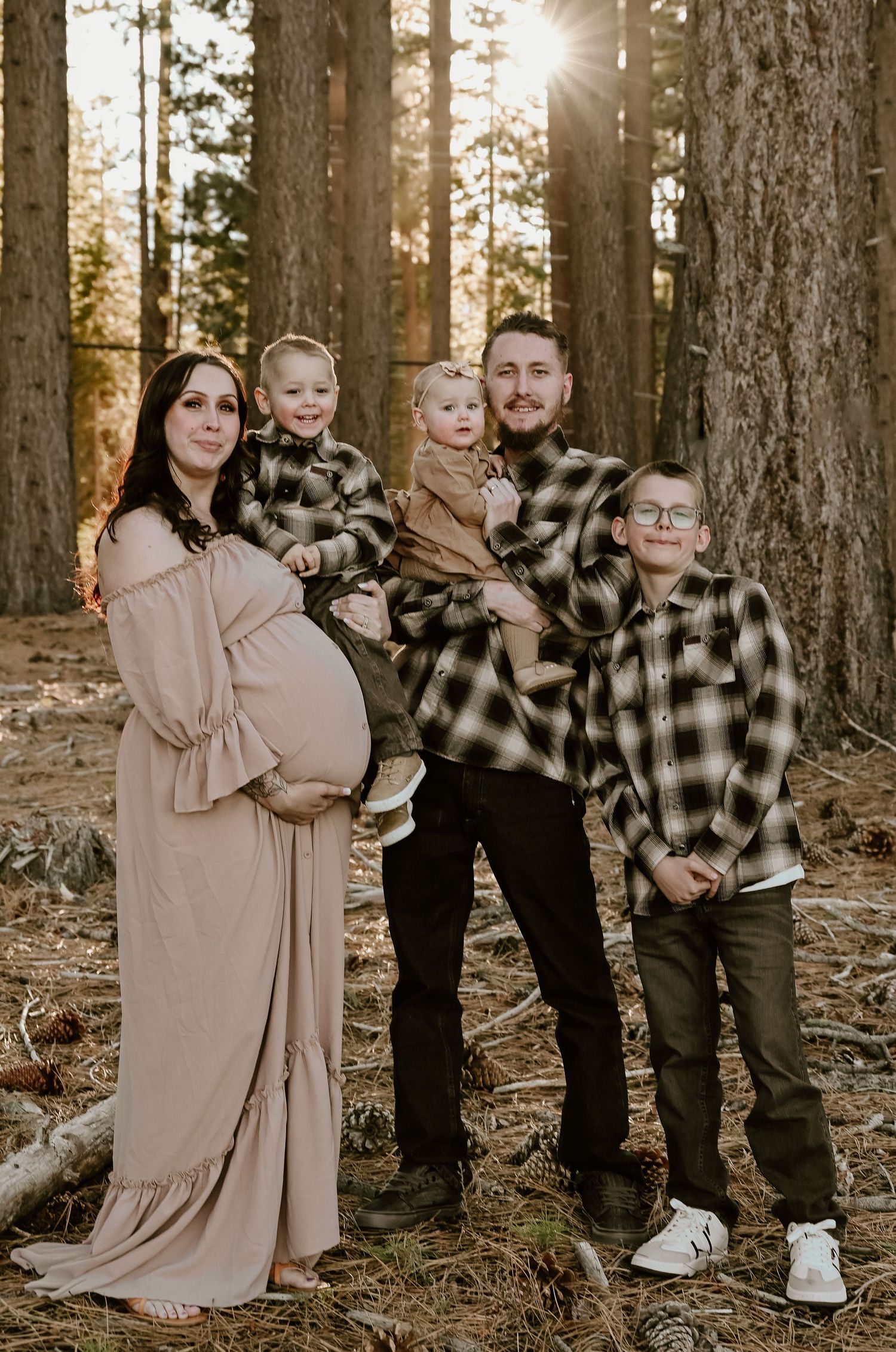 A family in plaid flannel and muted tones poses together in a sunlit forest setting during golden hour.