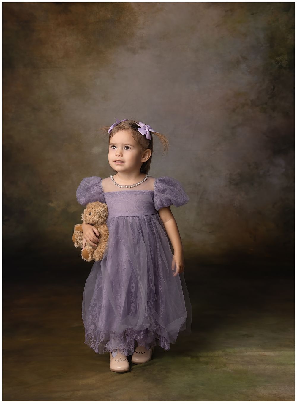 adorable fine art portrait of little girl, toddler, in purple tulle dress holding a teddy bear on a dark background