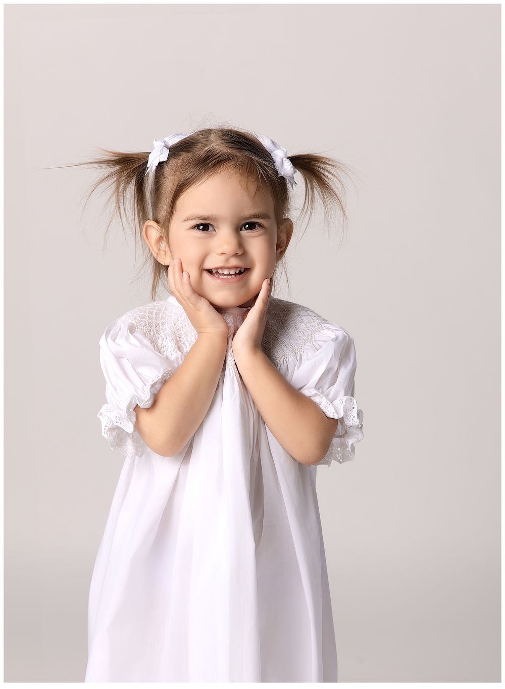 2 year session of adorable girl in white dress hands on chin smiling big with pigtails.