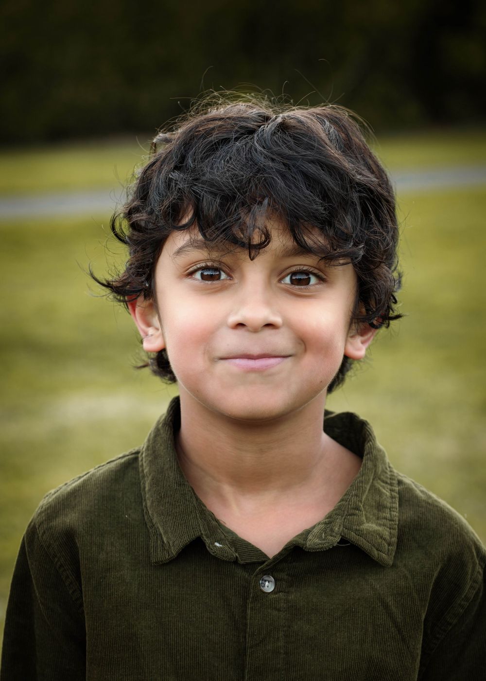 Portrait of a child wearing a dark green corduroy shirt with curly dark hair and gentle smile outdoors in soft natural light.