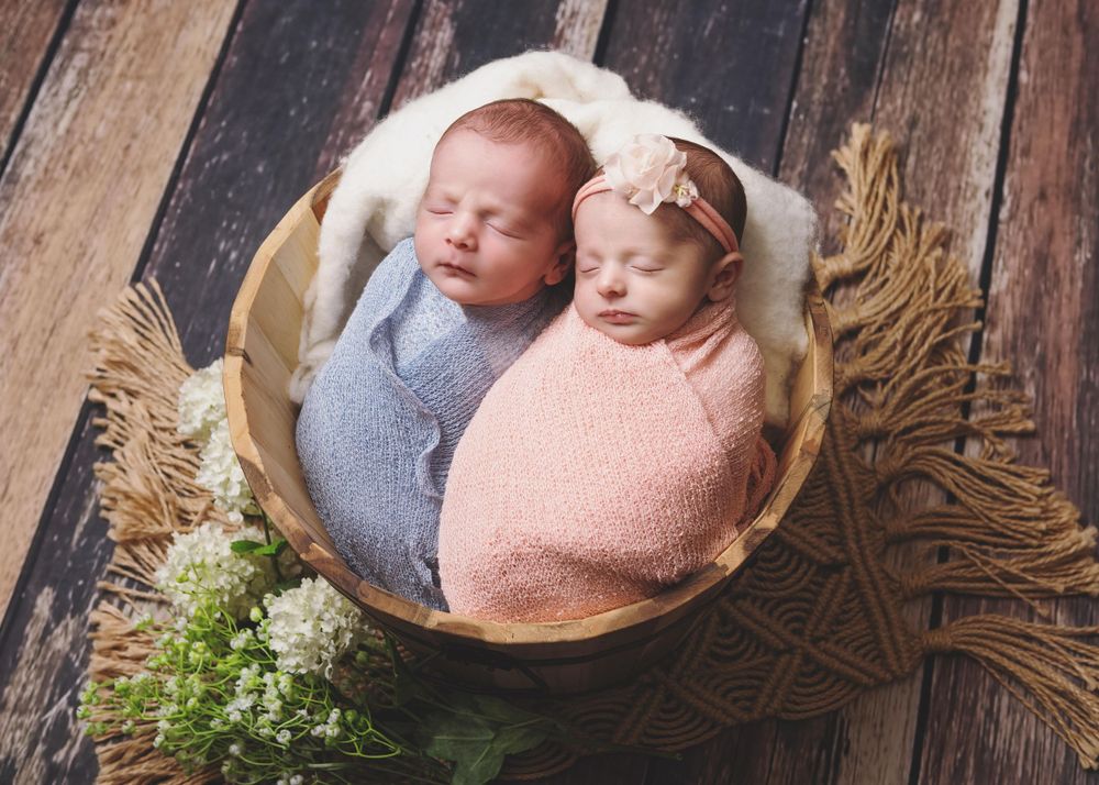 Twin newborns swaddled in pink and blue sleeping together in wooden bowl.