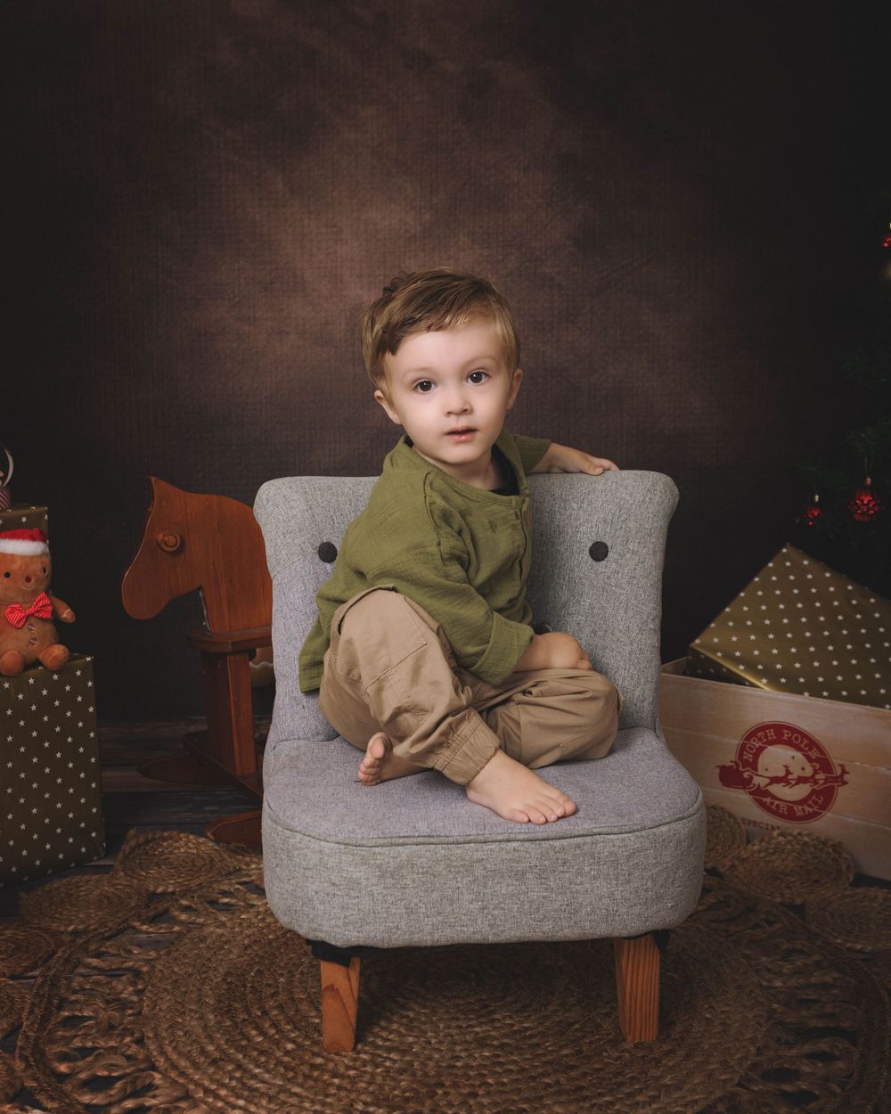 Small child sitting barefoot on a grey chair with toys and decorations in the background.
