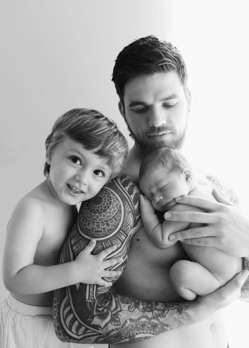 Black and white portrait showing tender moments between family with tribal tattoo and baby in natural lighting.