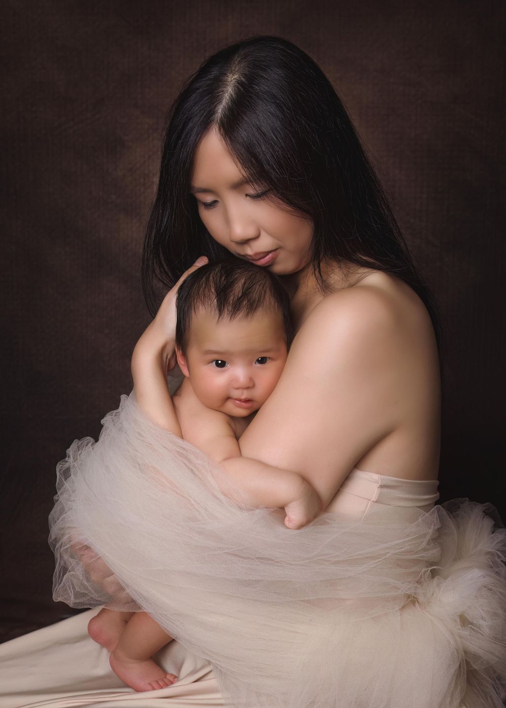 Intimate studio portrait of mother and baby draped in soft white tulle against dark background.