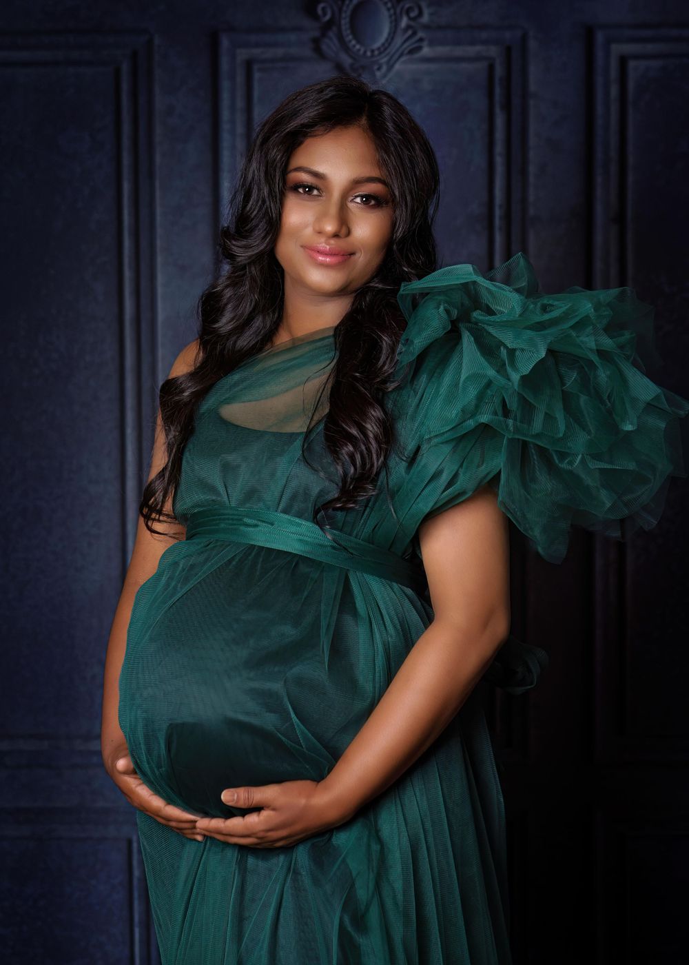 Maternity portrait featuring emerald green gown against dramatic dark blue wall.