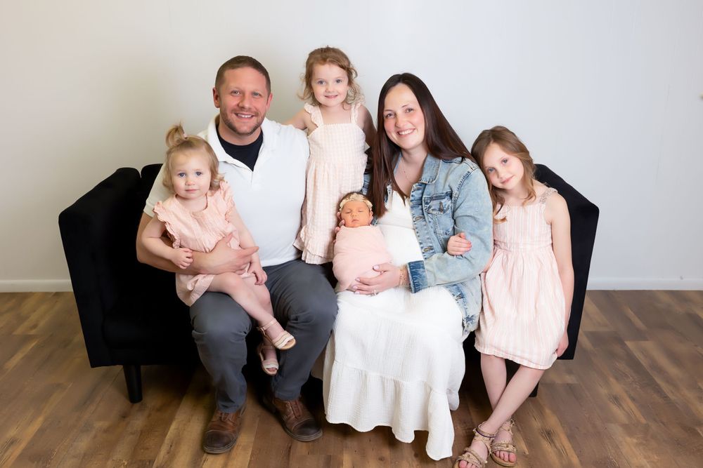 A family of six sits together on a black couch in a studio setting with wooden floors and light gray walls.