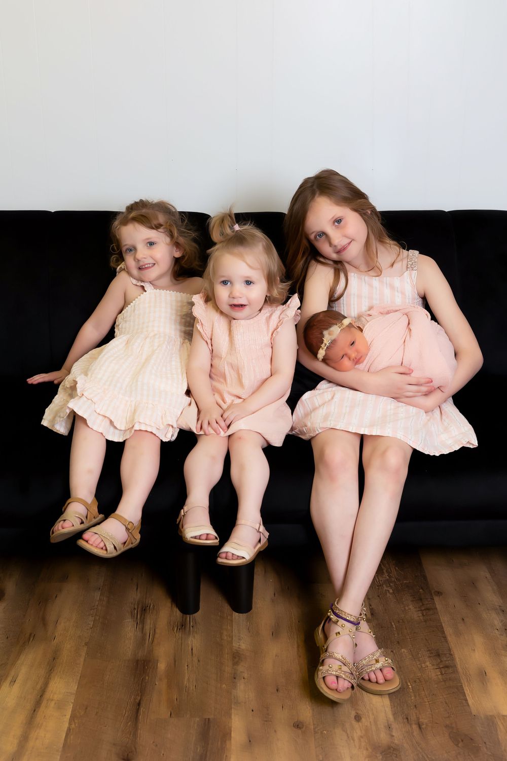 Four children wearing matching pink dresses sit together on a black couch in a studio setting.