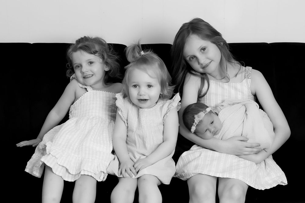Black and white portrait of four siblings in white dresses sitting together on dark furniture.