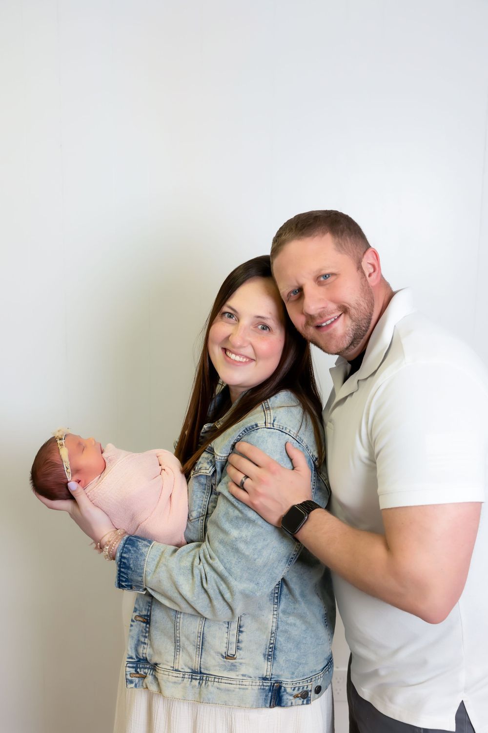 Couple in casual attire standing against white wall holding their newborn baby in pink clothing.