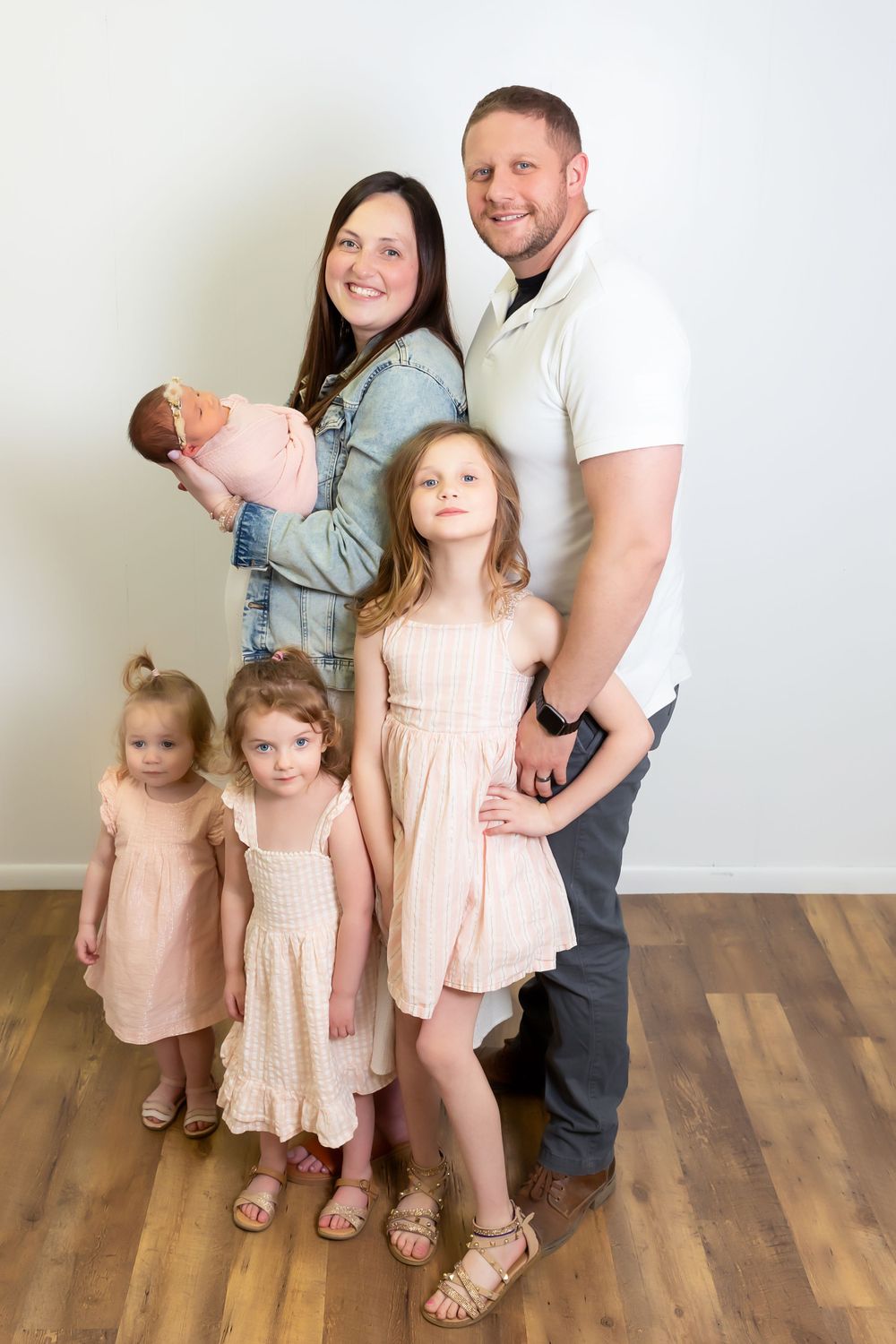 Family portrait with parents and four children in matching pink and neutral outfits standing on hardwood floor.