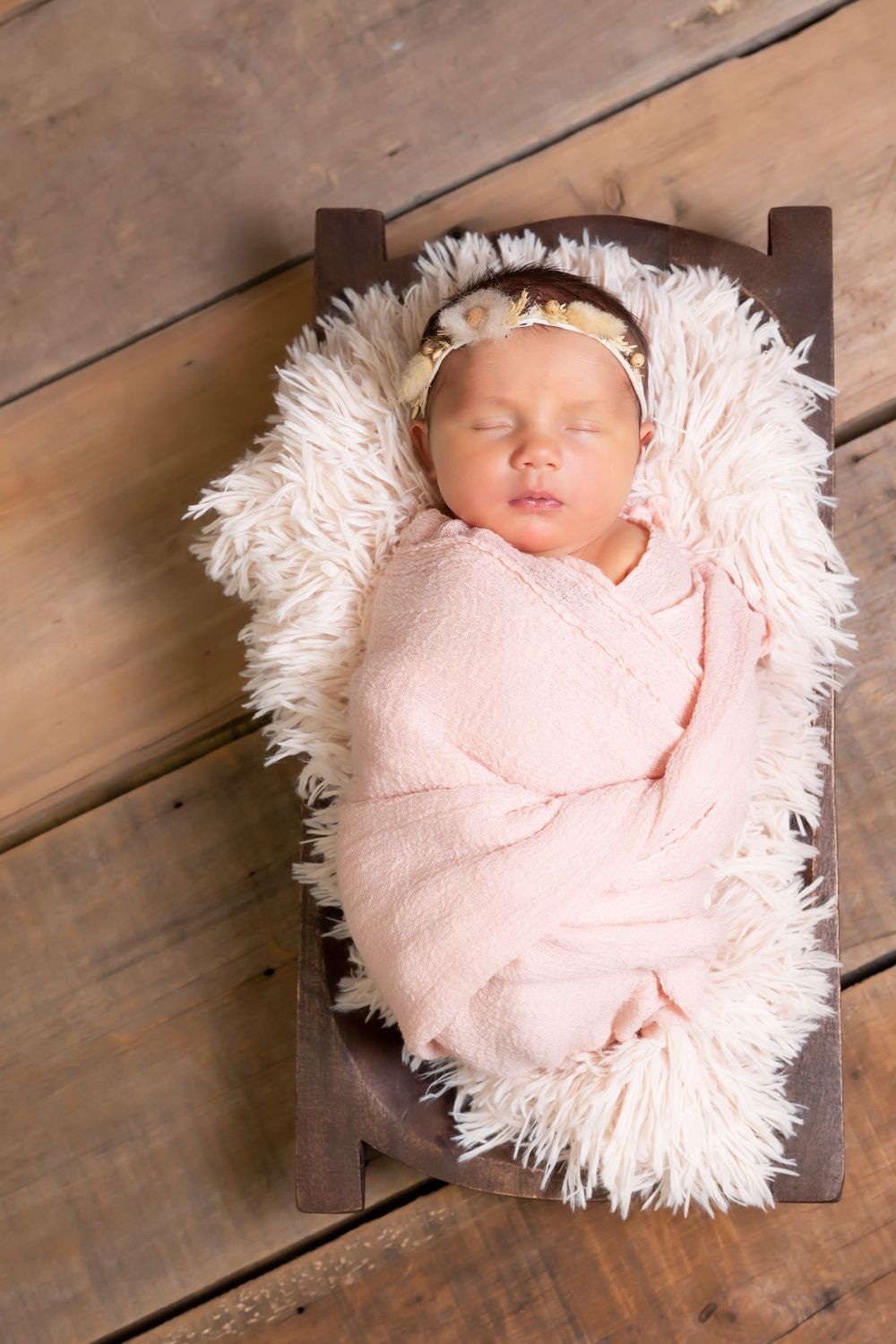 A sleeping newborn swaddled in soft pink blanket rests on white fur in a small wooden bed on wooden floors.
