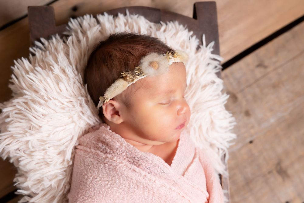 A sleeping newborn wrapped in soft pink swaddle lies on fluffy white fur with a delicate lace headband.