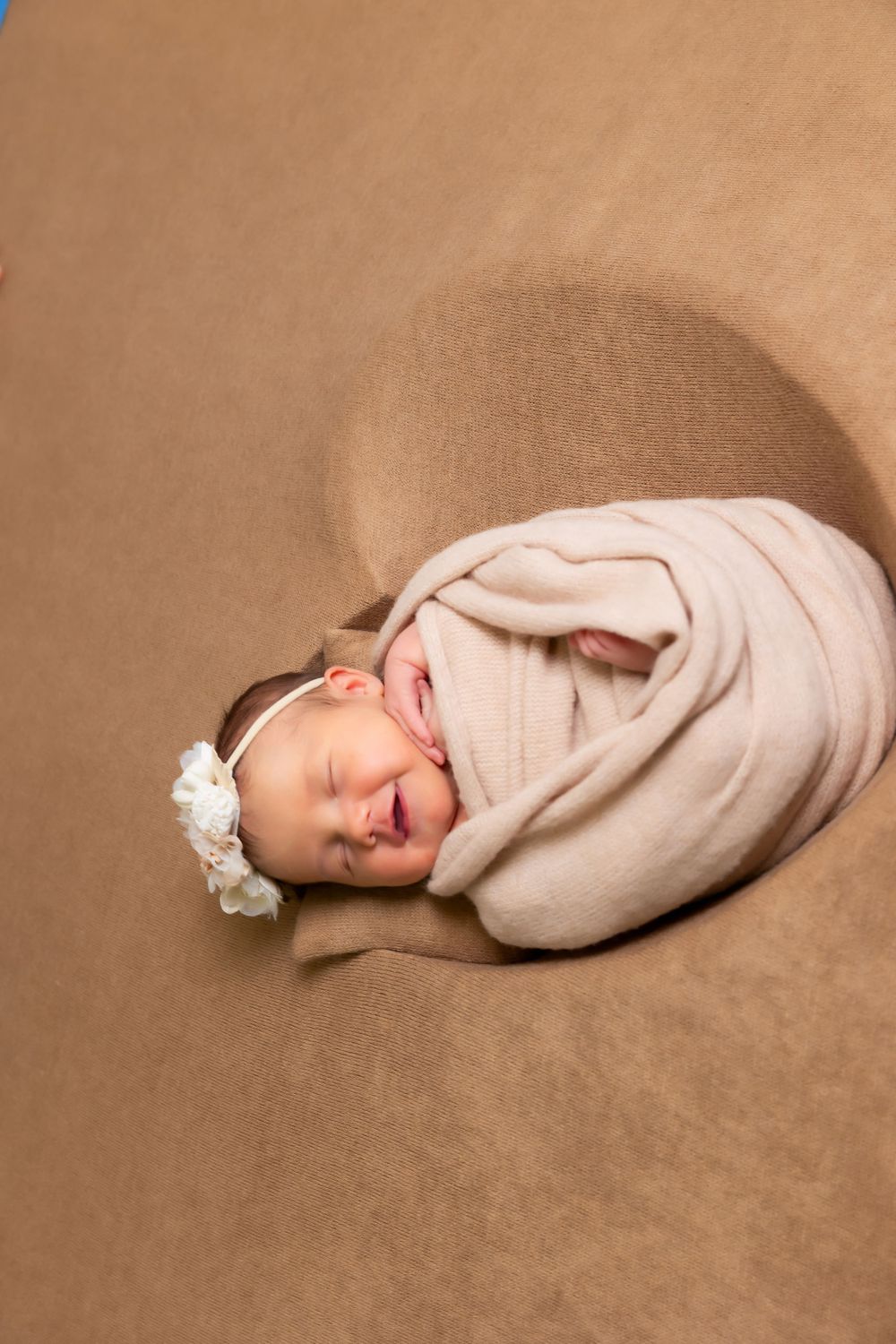 A sleeping newborn swaddled in beige fabric with a white headband lies curled up on a brown background.
