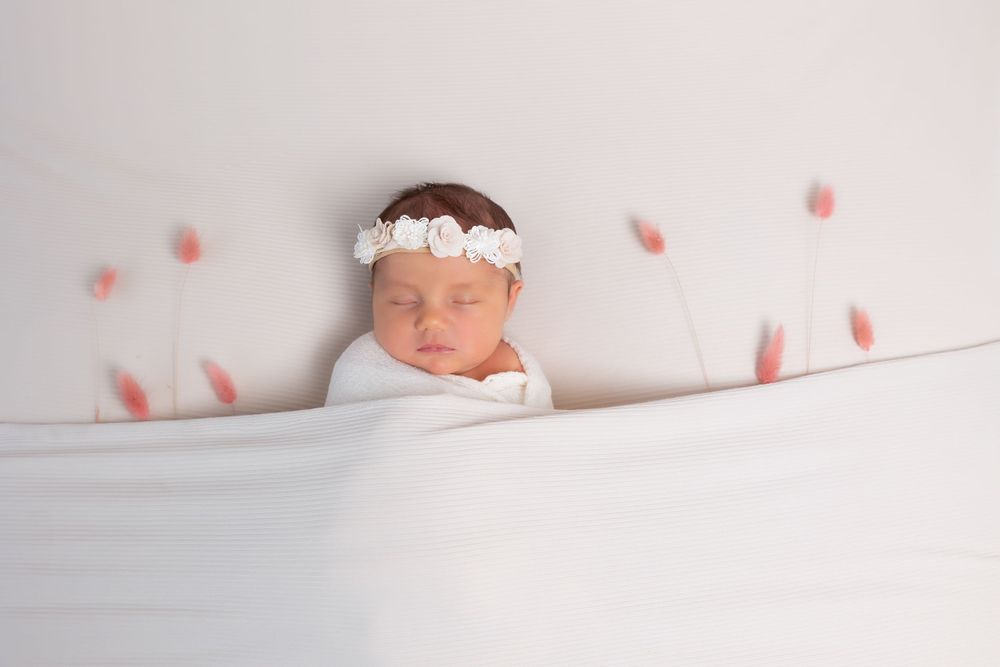 Sleeping newborn wrapped in white swaddle with floral headband against soft background with pink petals scattered around.
